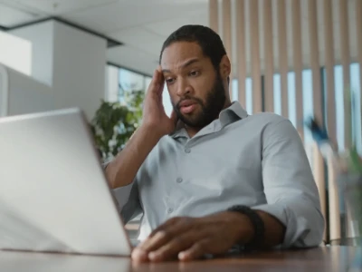  Ein Mann schaut auf seinen Laptop und hält sich dabei die Hand an die Stirn. Er hat Kopfschmerzen.