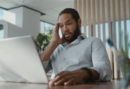  Ein Mann schaut auf seinen Laptop und hält sich dabei die Hand an die Stirn. Er hat Kopfschmerzen.
